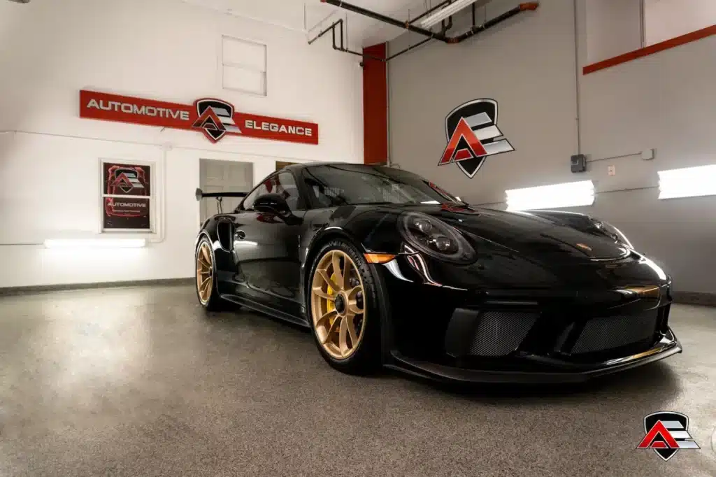 A shiny black Porsche GT3 with gold wheels, parked in a garage with Automotive Elegance signage.