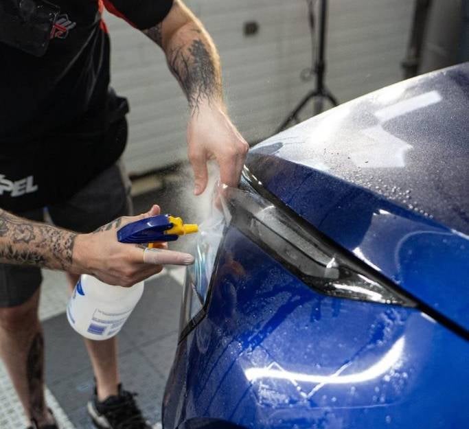 A tattooed person sprays liquid onto a car's blue hood while applying a clear protective film over the headlight.