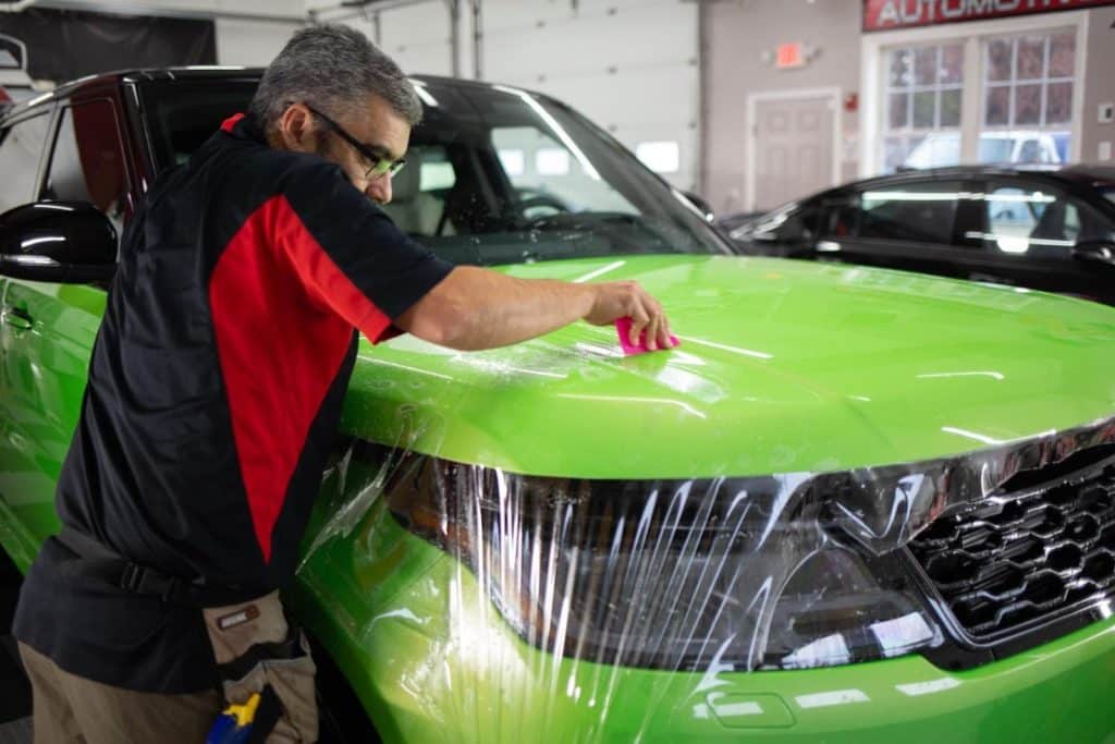 A man in glasses wearing a black and red polo shirt applies a protective vinyl wrap to the hood of a neon green car.