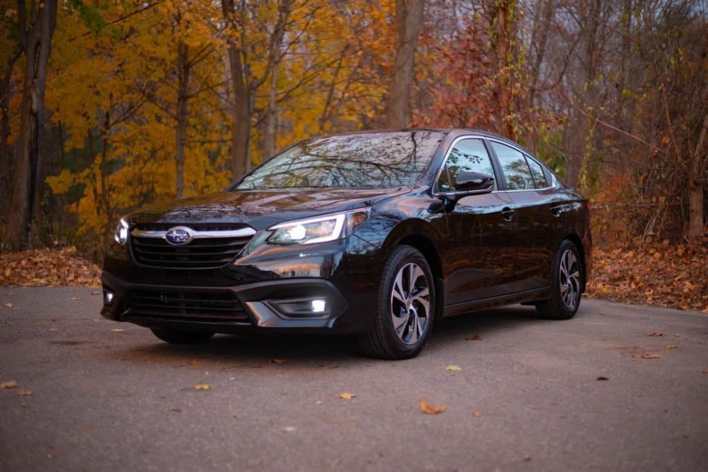 A shiny sedan is parked on an asphalt road with glowing headlights, autumn trees in the background.