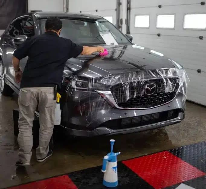 Man applying clear protective film to the hood and front of a Mazda CX-5 in a garage.