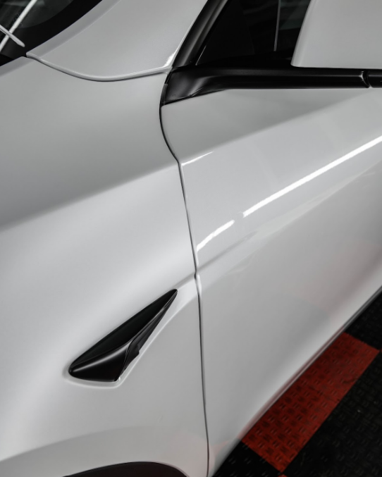 Close-up of a white car's front fender, door, and side mirror base, with a black trim accent near the wheel.