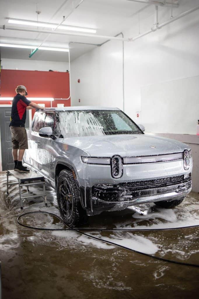 Man washing a silver Rivian electric SUV, covered in white soap suds, on a wet concrete floor.
