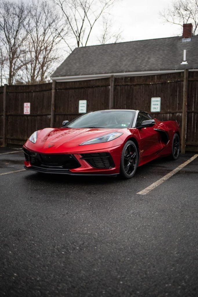 Red Chevrolet Corvette C8 parked in an asphalt lot with a dark wooden fence and building in the background.