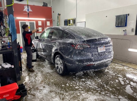 A person in a garage sprays soap onto a dark gray Tesla, creating foam covering the car and floor.
