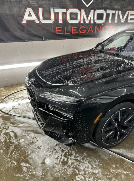 Front corner of a black car, covered in white soap suds, being washed in front of an Automotive Elegance sign.