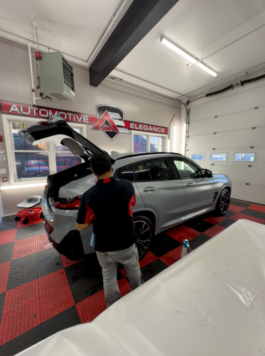 A man in a garage with red and black checkered floors works on the open trunk of a gray BMW SUV.