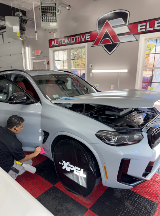 A man applies a protective film to the side of a light gray car with an open hood in a garage.