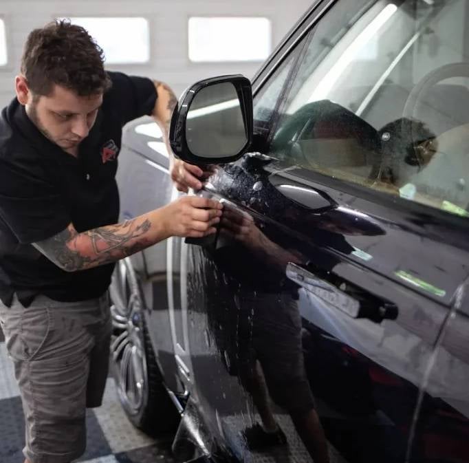 A man with tattoos on his arms applies a protective film to the side of a black car.