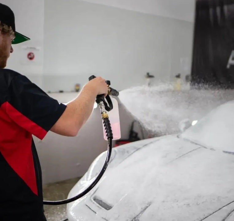 A man in a black and red shirt spraying a white sports car with thick white foam from a hose attachment.