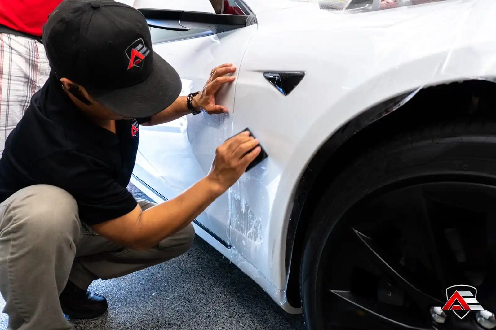 Technician applying a clear protective film onto the side panel of a white car, smoothing it with a tool.