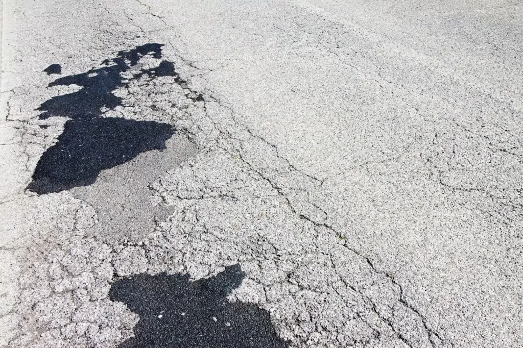 Severely cracked light gray asphalt with dark gray patches and a dark, irregular spill.