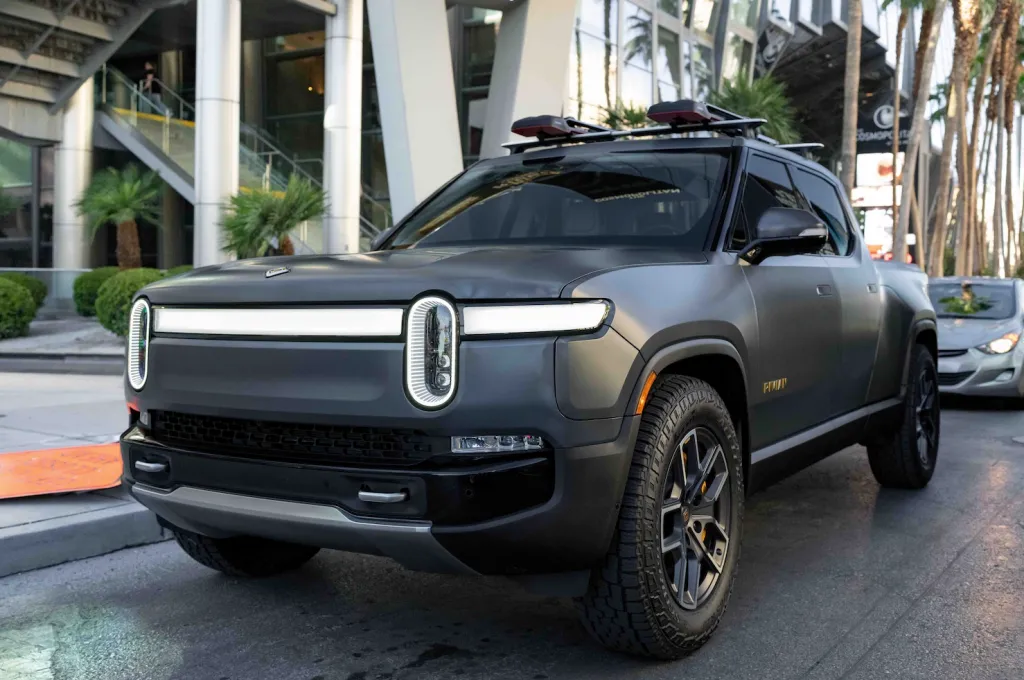 A matte grey Rivian R1T electric pickup truck with a roof rack and light bar, parked on a city street.