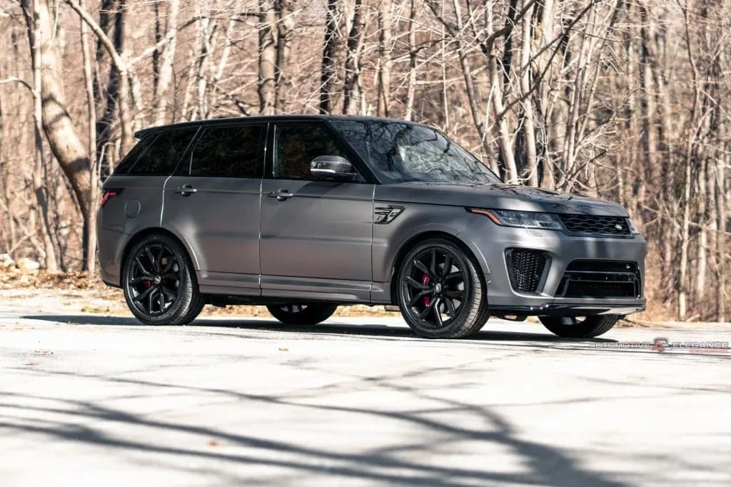 A matte dark gray Range Rover Sport with black wheels and red brake calipers parked on pavement with trees in the background.