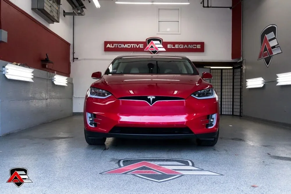 Front view of a shiny red Tesla Model X in a garage with "Automotive Elegance" signage and logos.