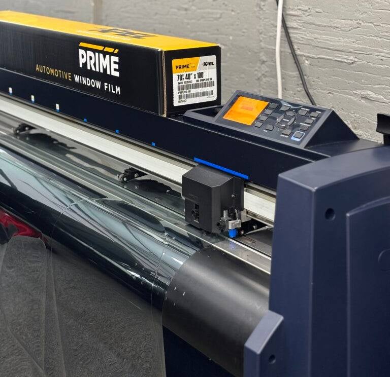 A large format plotter cutting Prime Automotive Window Film from a roll of dark, reflective material.