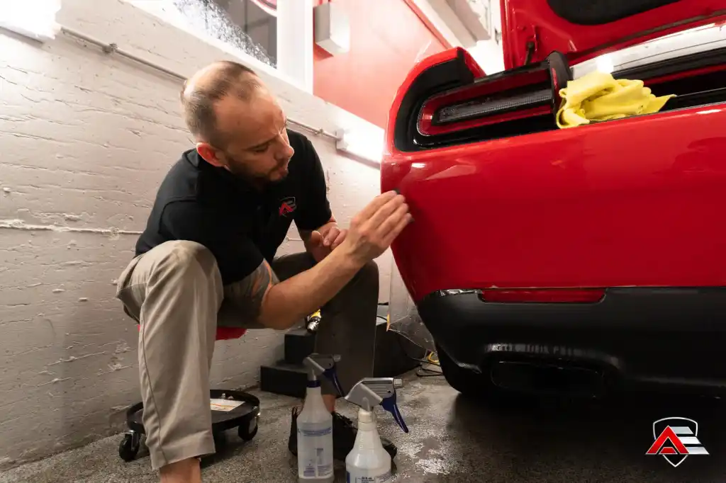 Technician inspecting paint protection film installation on the rear bumper of a red Dodge