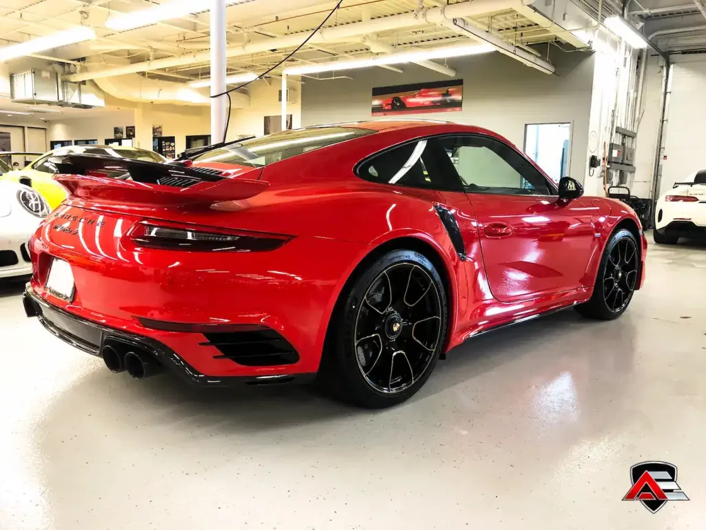 Rear 3/4 view of a shiny red Porsche 911 Turbo S parked on a showroom floor with other cars in the background.
