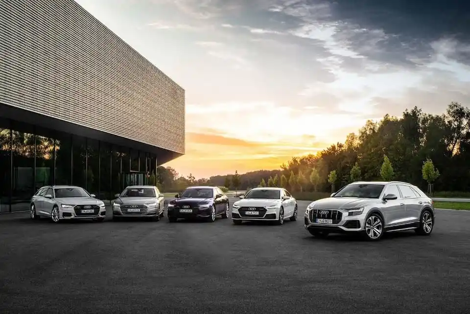 Five Audi cars parked in a row on asphalt, with a modern building and trees in the background at sunset.