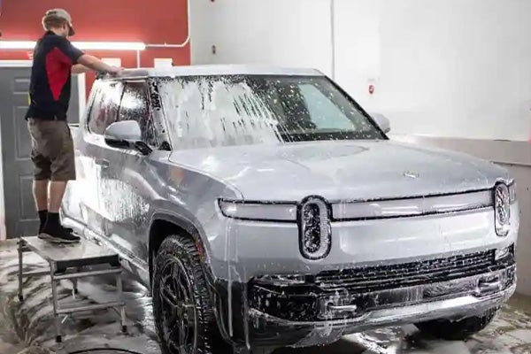 A person standing on a stool washes a foamy silver Rivian R1T pickup truck.