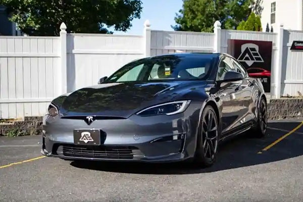 A dark gray Tesla Model S parked on asphalt in front of a white picket fence and a business sign.