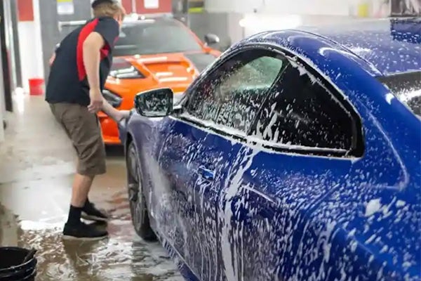 A man in a garage washes a blue sports car covered in white soap suds, with an orange car in the background.