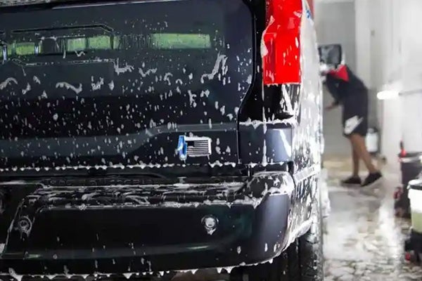 Close-up of a dark SUV's rear covered in white soap suds, with a person washing it in the background.