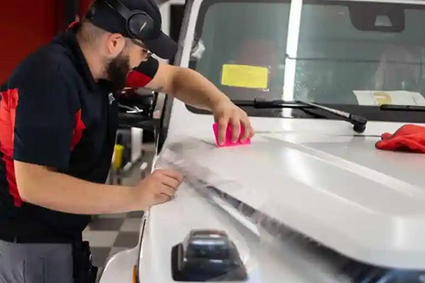 A man in headphones and a cap applies a clear protective film to the hood of a white vehicle.