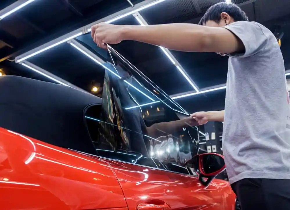 A person applies window tint to the side window of a glossy red convertible car.