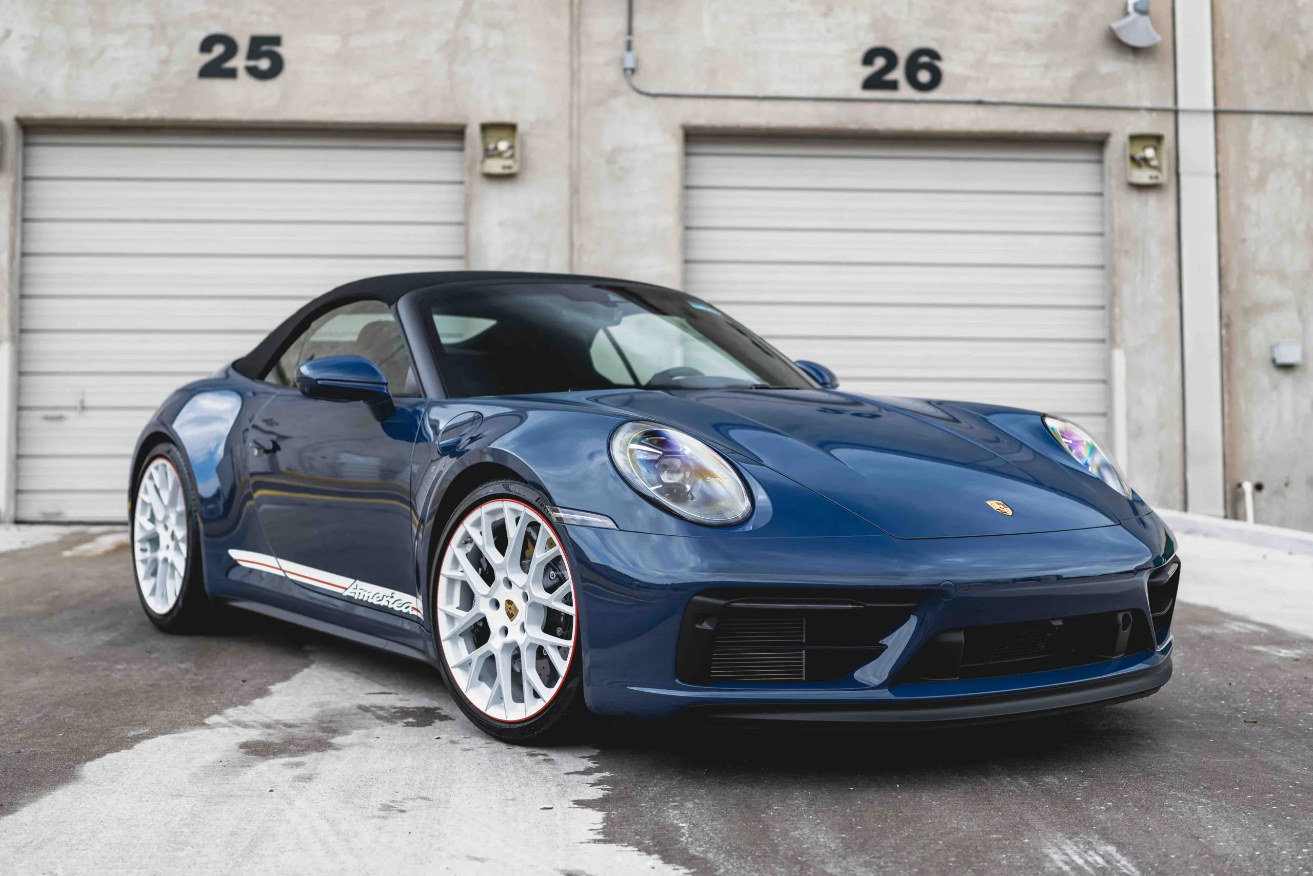 A dark blue Porsche 911 convertible, with white wheels and red trim, parked before two garage doors.