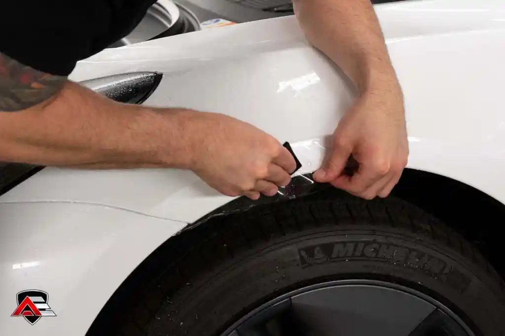 Two hands apply protective film to the white car's fender near the tire, using a squeegee to smooth it.