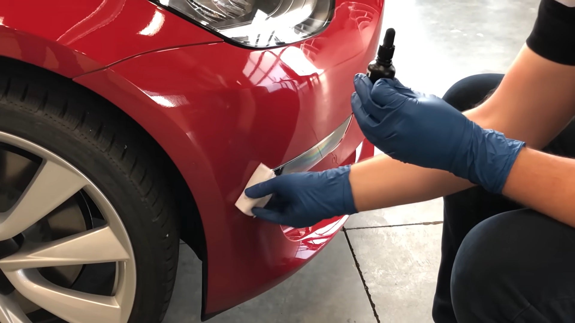 Person in blue gloves applies a coating with a cloth to a shiny red car bumper next to a tire.