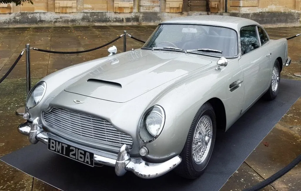 A silver Aston Martin DB5, similar to James Bond's car, parked outdoors with its distinctive front grille visible.