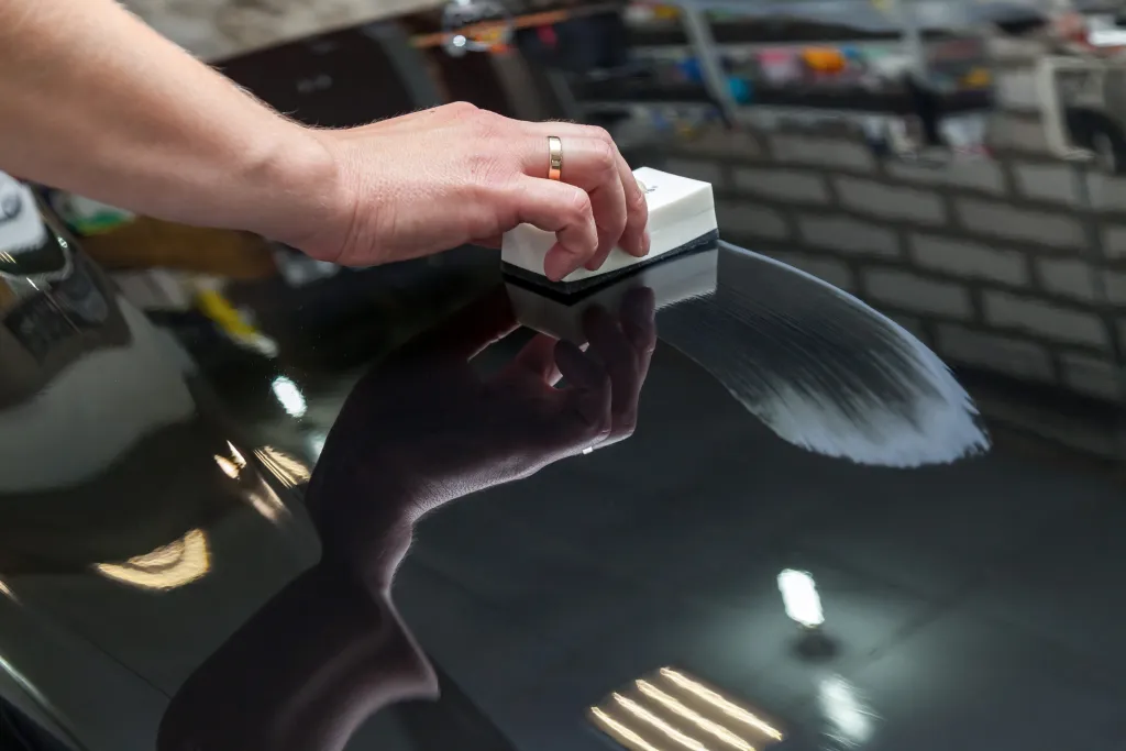 Hand with a ring applying a clear coating to a glossy dark surface with a white applicator, leaving a distinct streak.