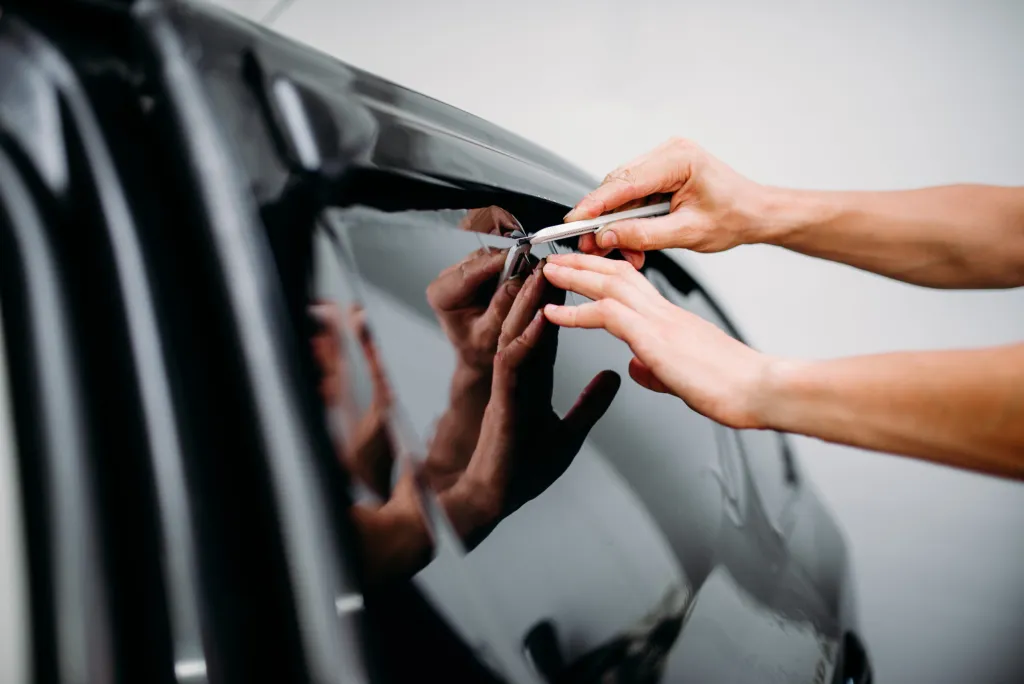 A close-up of hands applying dark window tint film to a car window with a utility knife.