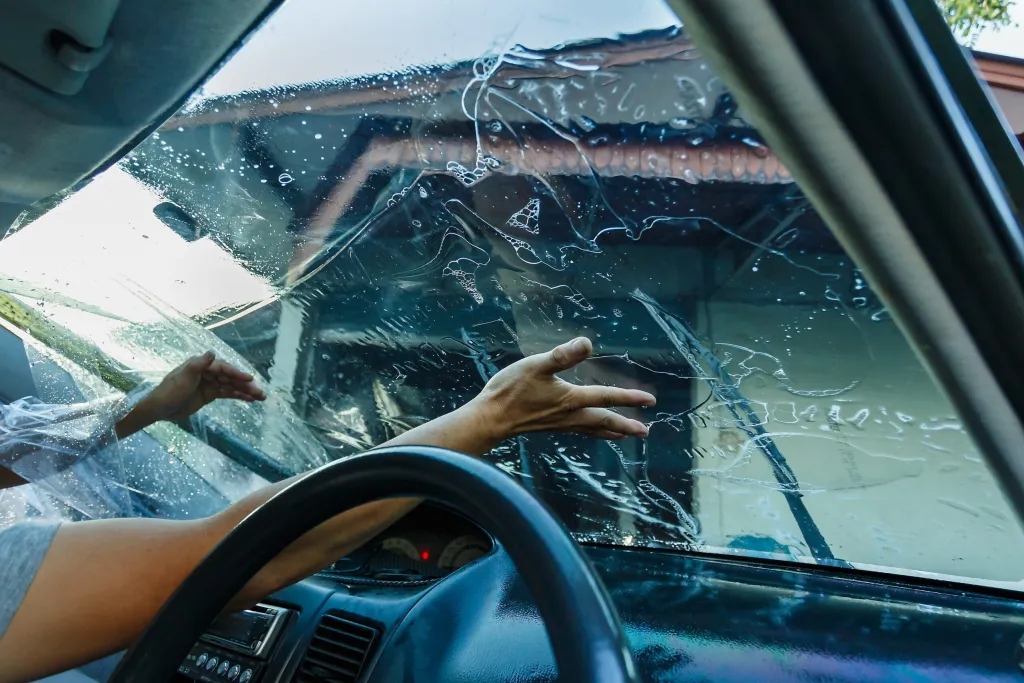 A person's hands applying clear plastic film, likely window tint, to a car's wet front windshield from the inside.