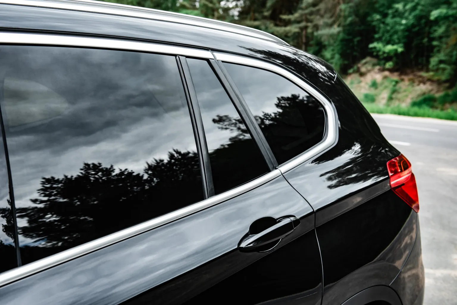 Side view of a glossy black car with tinted windows reflecting trees and a cloudy sky.