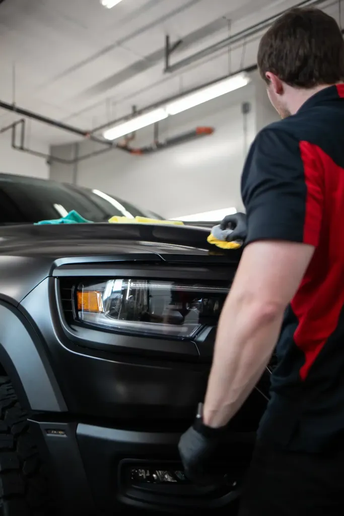 Man in black and red polo shirt with gloves polishing a matte black truck's hood and headlight.