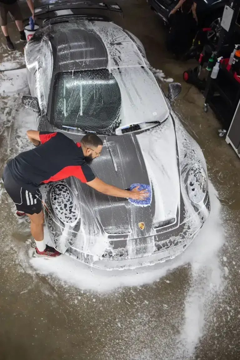 Technician hand-washing a Porsche sports car covered in foam before paint protection film installation