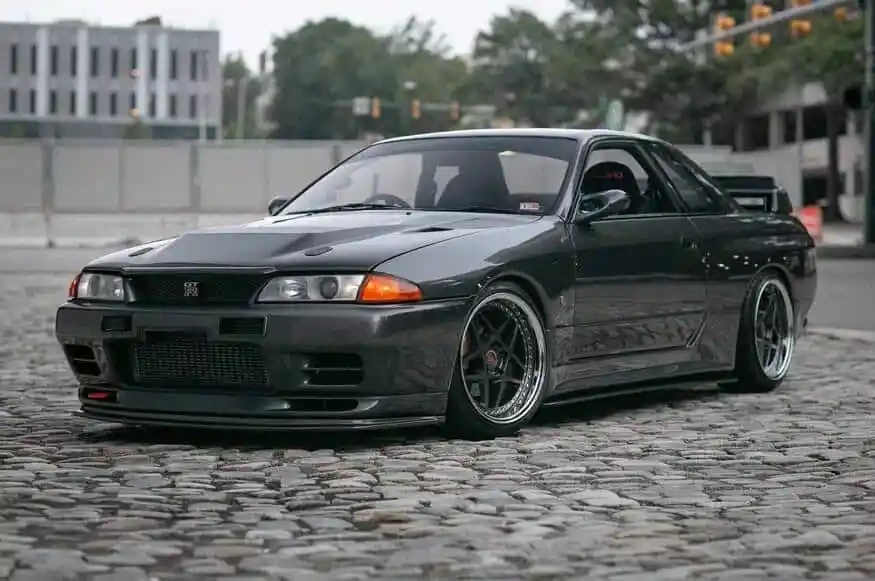 A dark grey Nissan Skyline GTR with a hood scoop, front lip, and aftermarket wheels on a cobblestone street.
