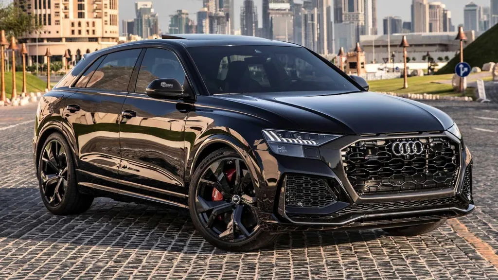 Black Audi RSQ8 SUV parked on a stone-paved street, with a city skyline in the background.