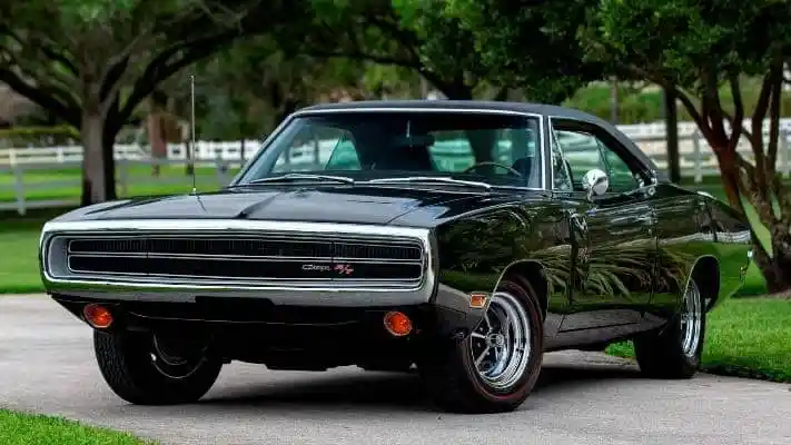 A black 1969 Dodge Charger R/T parked on an asphalt driveway with trees and a white fence in the background.