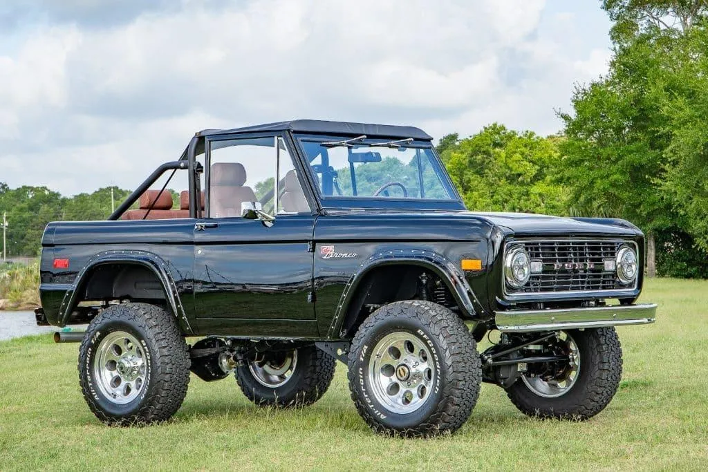 A restored black early Ford Bronco with a soft top, roll bar, lifted suspension, and large off-road tires, parked on grass.