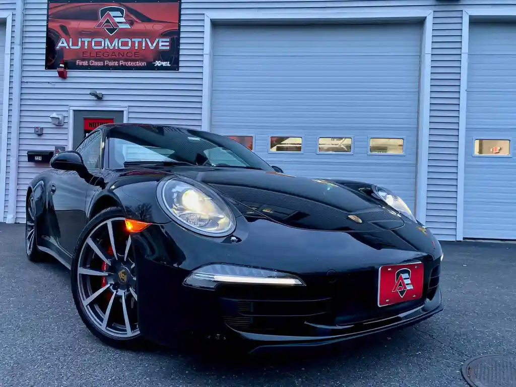 A shiny black Porsche sports car with red custom license plates parked outside a garage with a red sign above.