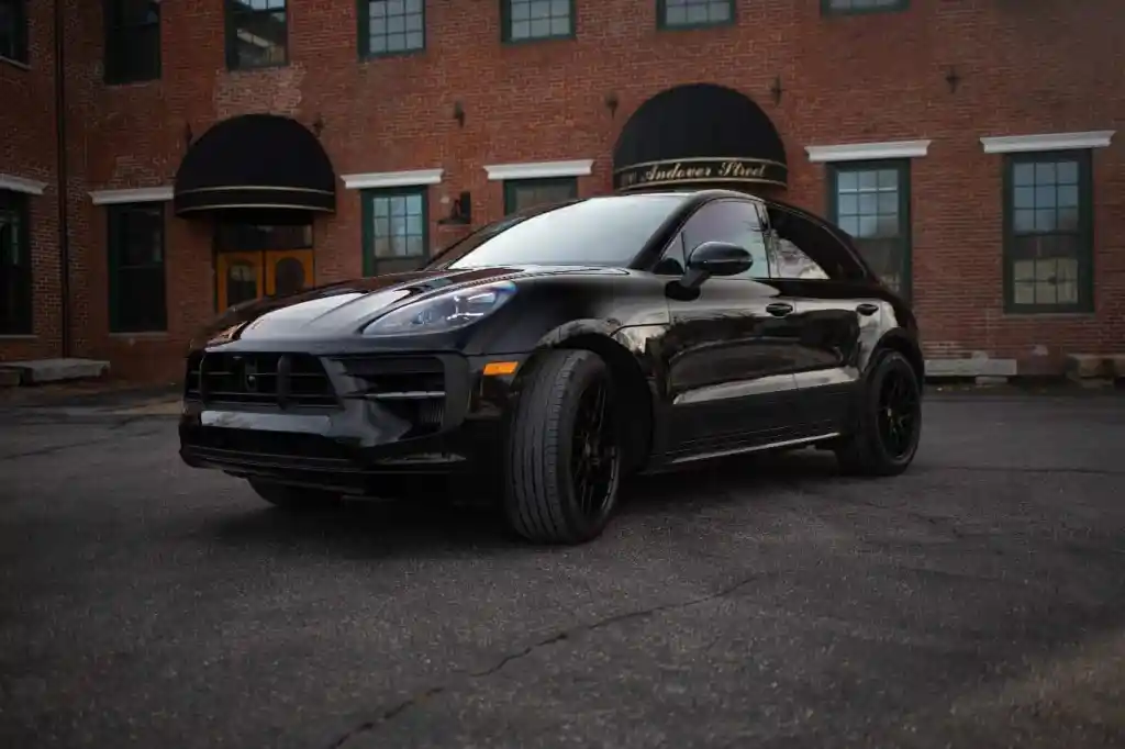A sleek black Porsche Macan parked on asphalt, angled slightly left, with a historic brick building in the background.