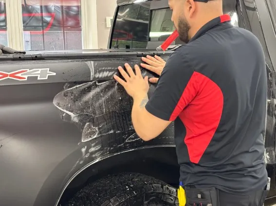 A man applies a protective film to the side of a black truck, with visible bubbles and liquid under the film.