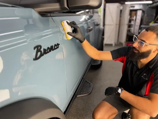 A bearded man in black gloves applies a ceramic coating to a light blue Ford Bronco.