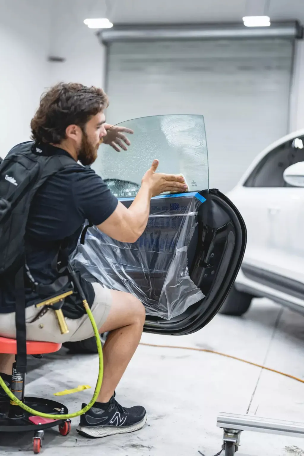 A man on a stool applies a protective film to a car window that's covered in suds, with the door exposed.