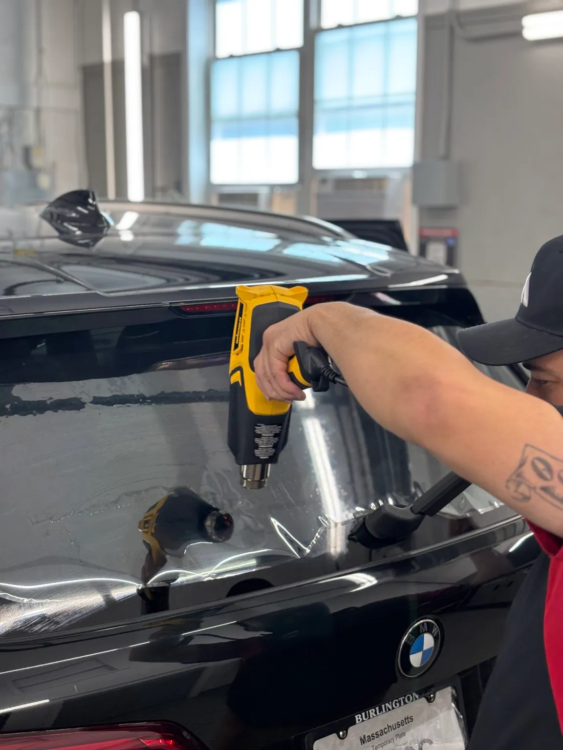 Person using a yellow heat gun to apply clear window film to the rear windshield of a black BMW SUV.