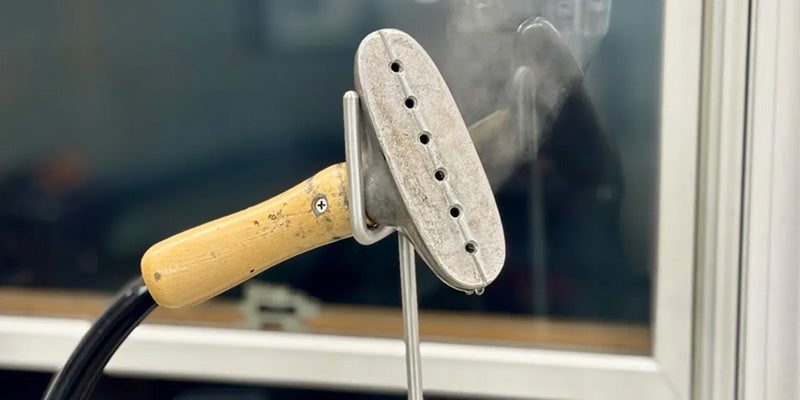 An old, handheld clothes steamer with a light wooden handle and a metal head emitting steam.